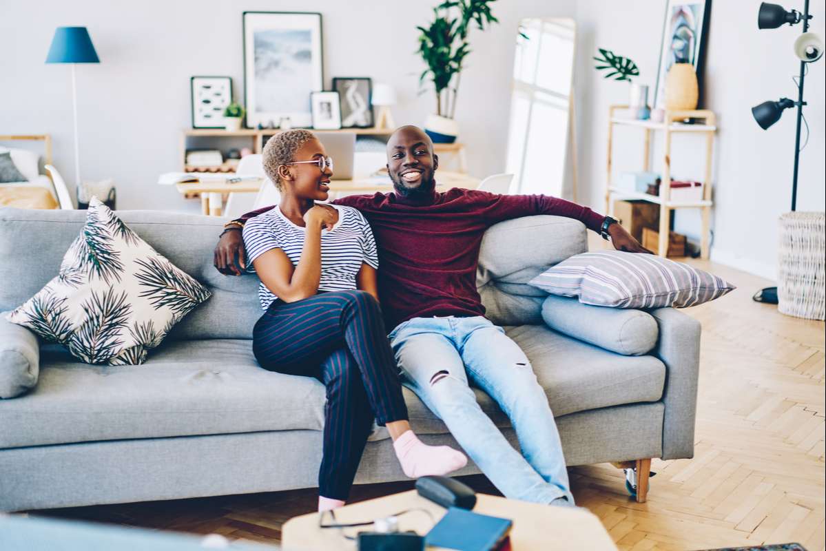 Beautiful dark skinned woman talking to smiling boyfriend sitting together on cozy sofa in living room (R) (S)